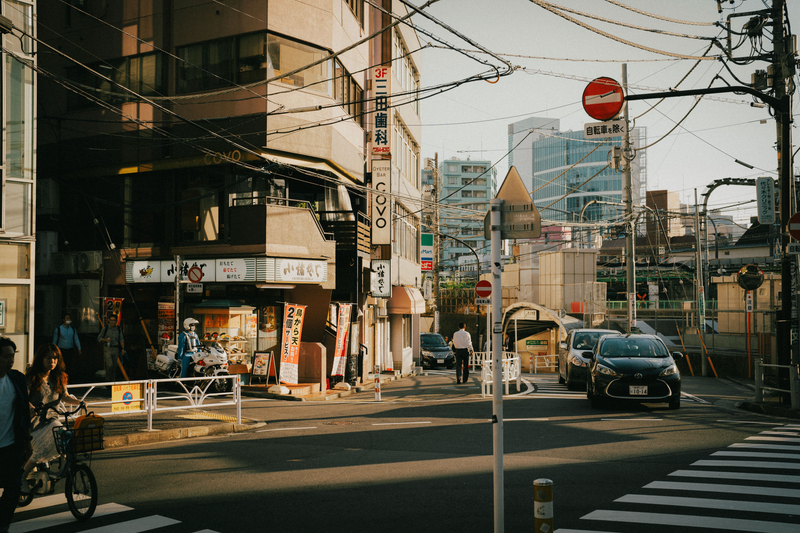 A narrow Tokyo street in late-afternoon light, with a police motorcyclist and a woman on a bicycle on the left, a black taxi-style car at the junction and overhead wires crossing the sky.