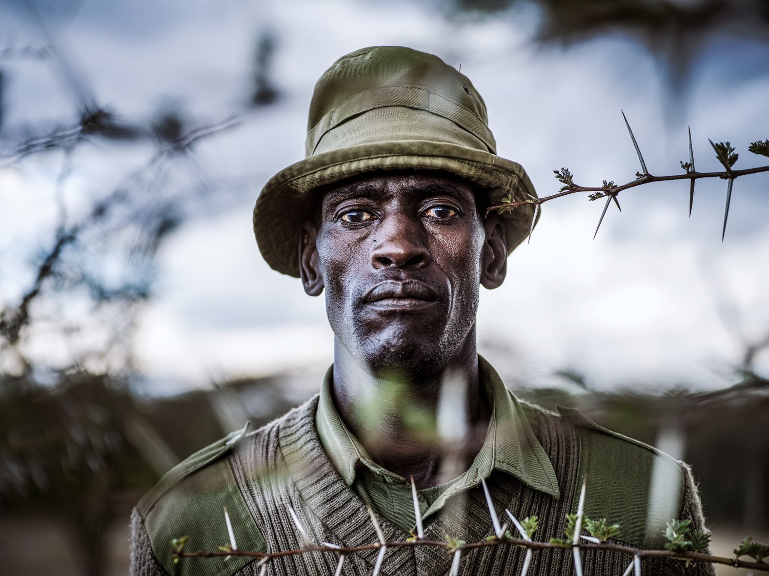 A ranger in a bush hat seen through a tangle of thorny acacia branches.