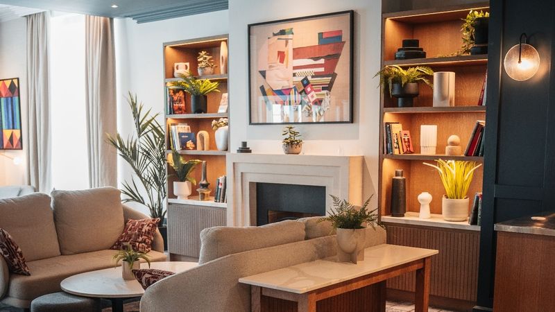 A hotel lounge with a cream sofa and curved armchair, a marble-topped console table, a white fireplace under a colourful abstract artwork, and lit wooden shelves holding plants, books and ceramics.