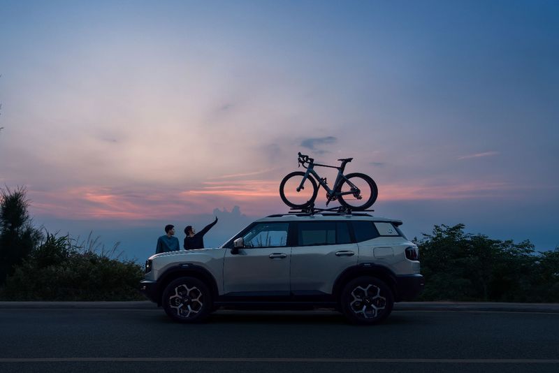 A white Jetour SUV with a road bike on its roof rack parked by a road at dusk as two people stand beside it under a pink and blue sky