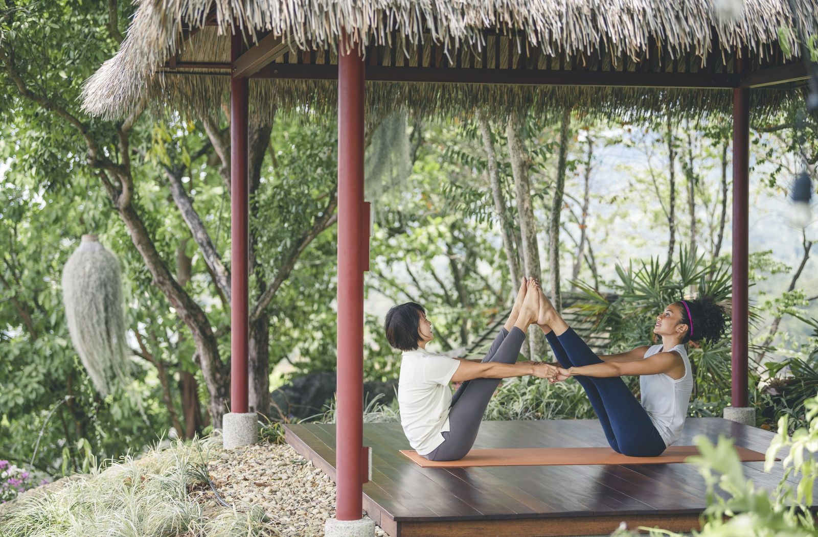 Two women do a partner yoga stretch on a mat in an open thatched pavilion surrounded by tropical trees.