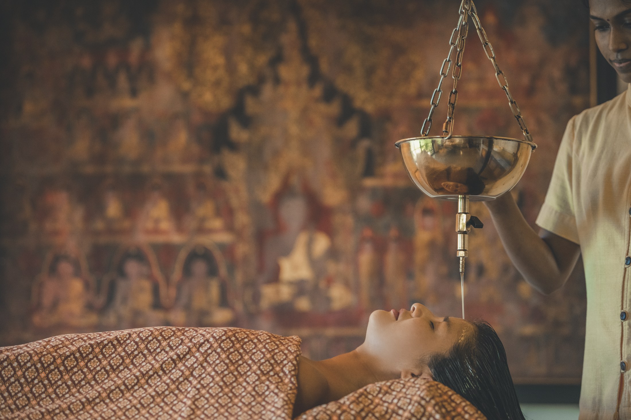 A woman lies under a patterned blanket as a therapist pours a thin stream of warm oil onto her forehead from a hanging brass vessel.