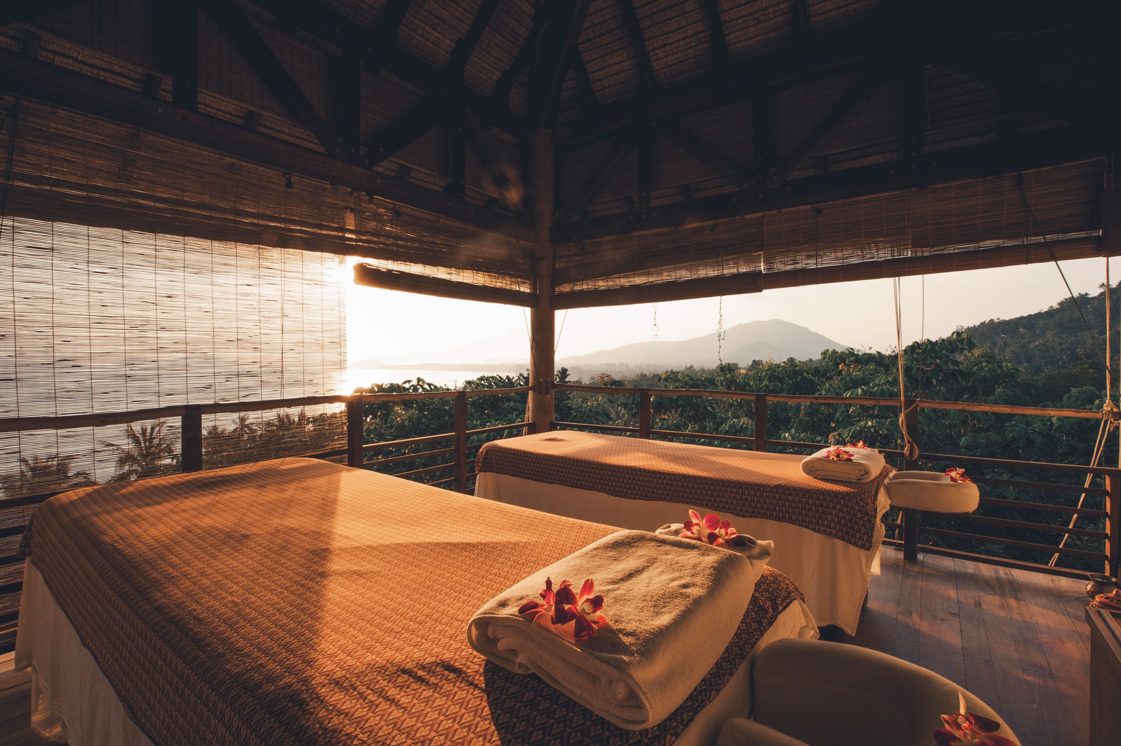 Two massage beds with folded towels and red flowers in an open timber treatment pavilion overlooking hills at sunset.