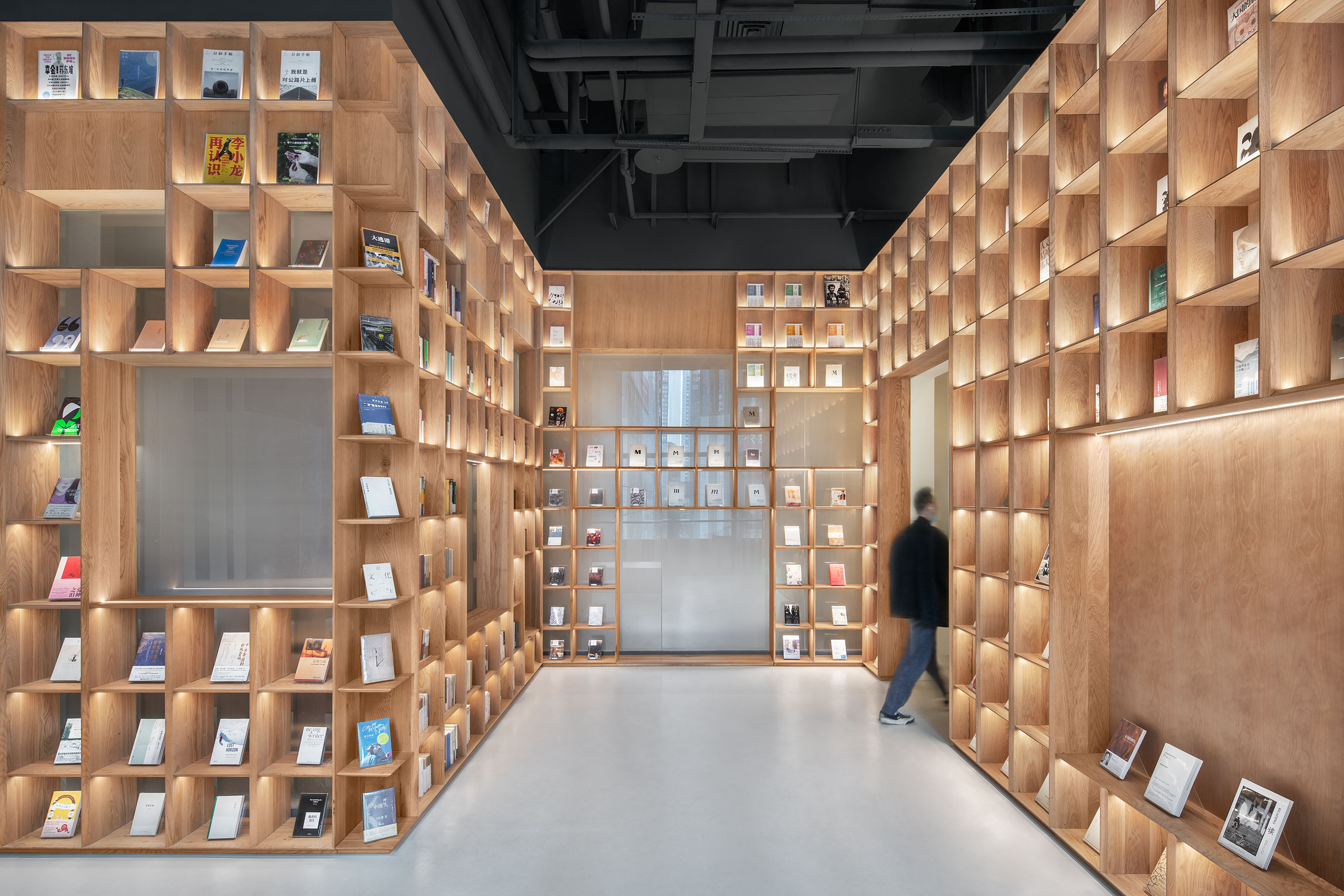 The Glade Bookstore: a narrow aisle lined with timber shelving cubes lit from within, with a person walking along it.