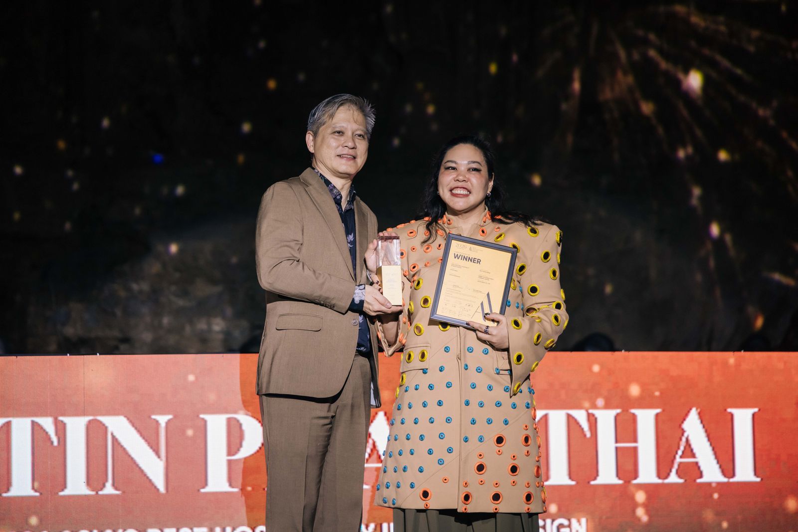 Two people on an awards stage hold a trophy and a “Winner” certificate in front of a lit backdrop.