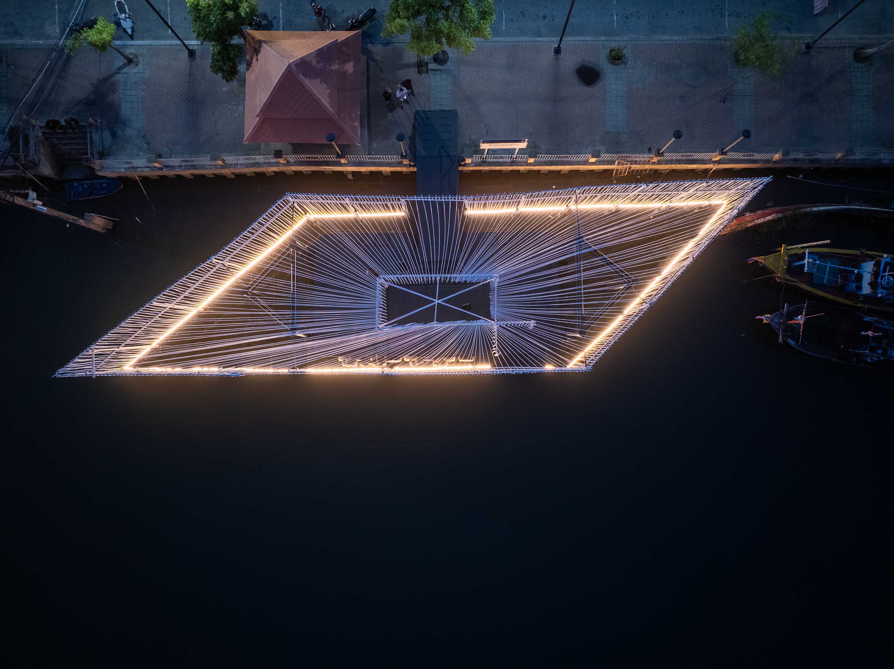Pattani Fishermen Community Hub: a night aerial view of a lit, diamond-shaped frame strung with lines over dark water, with fishing boats and a small shelter at the edge.