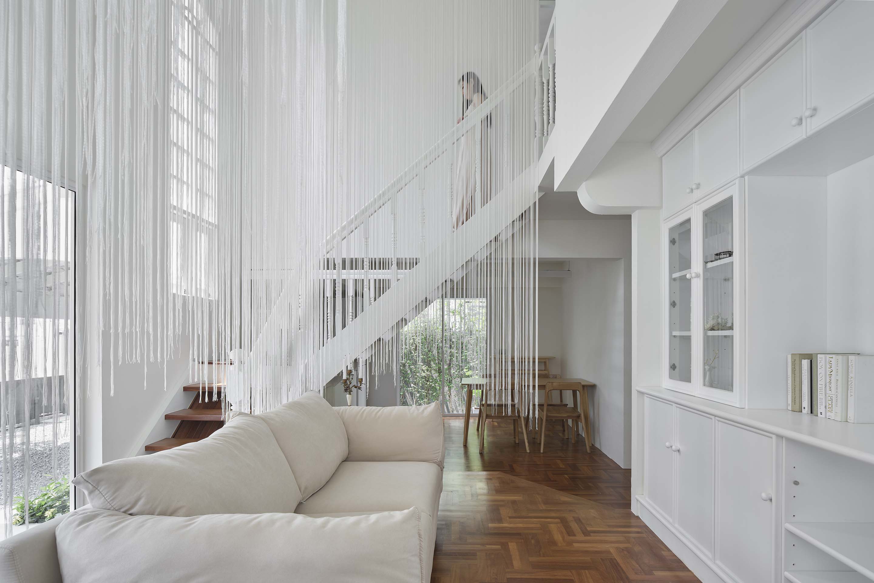 InJoy Snow Hotel Bangkok: a white double-height interior with hanging white strands, a staircase, a cream sofa and built-in cabinets above a parquet floor.