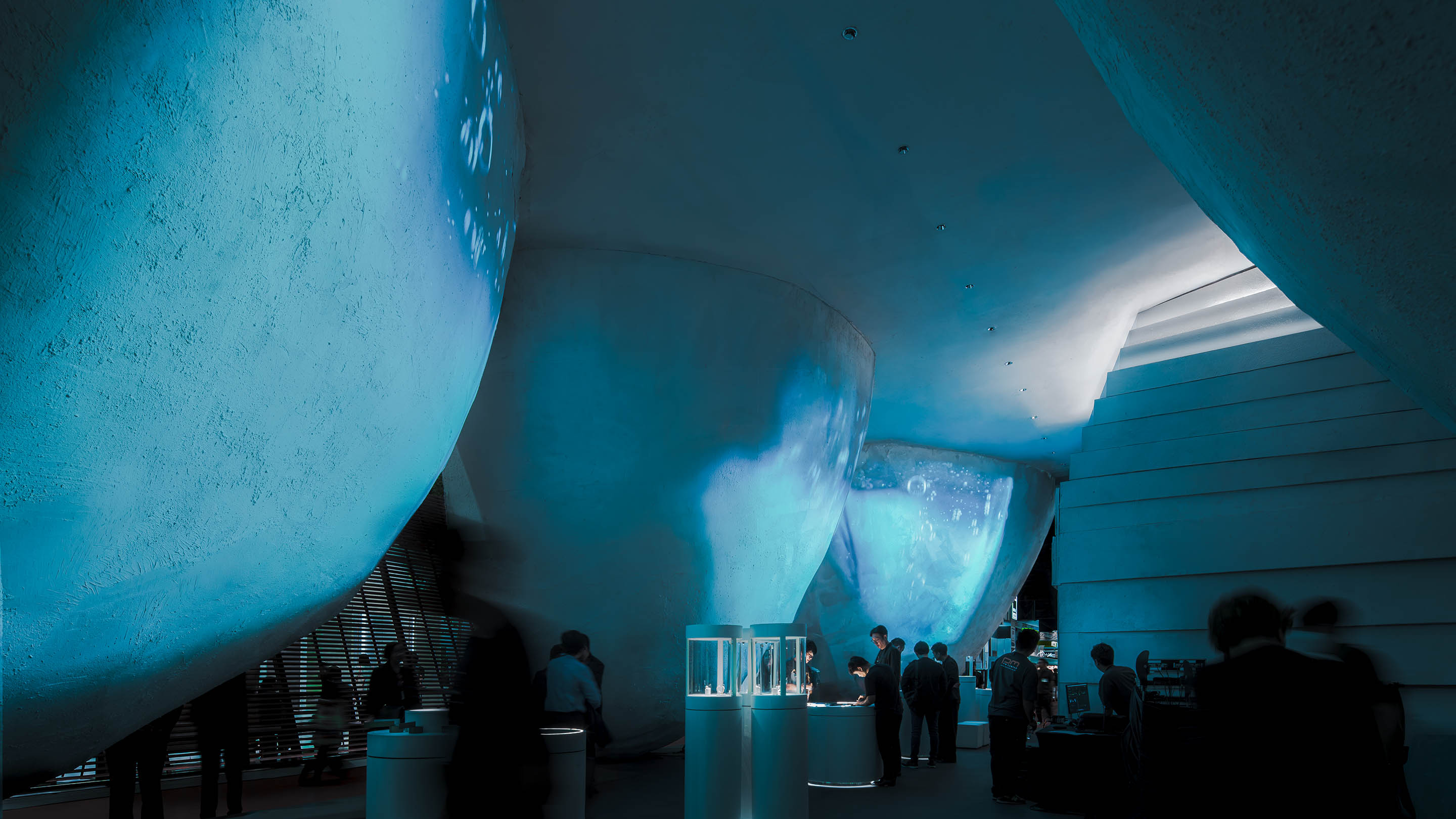 Mega Hill and Common Earth: visitors in a dim, blue-lit gallery beneath large curved white forms, with glass display cases in the foreground.