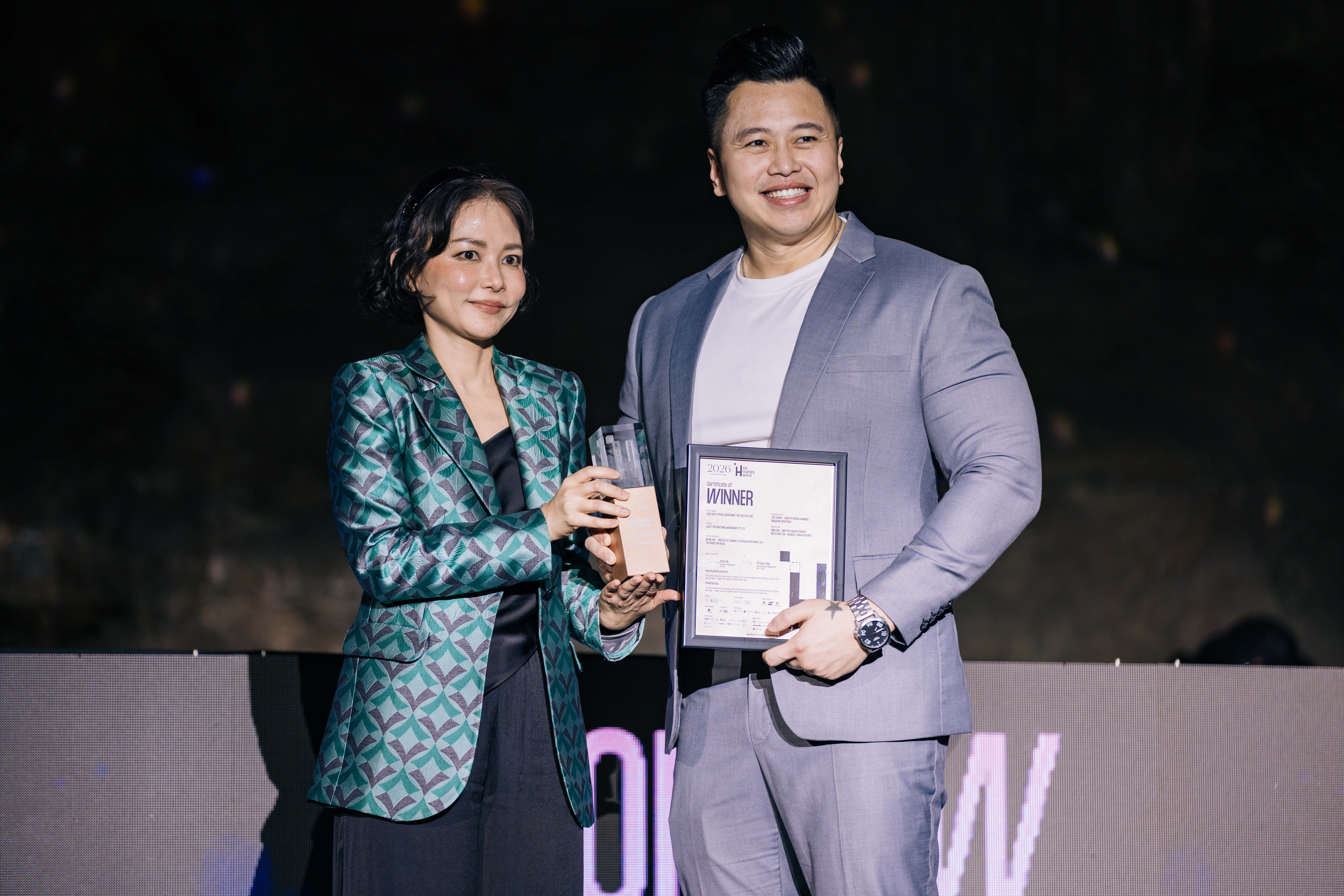 Aticha Chareerat, in a teal patterned blazer, holds a trophy beside a man holding a “Winner” certificate on stage.
