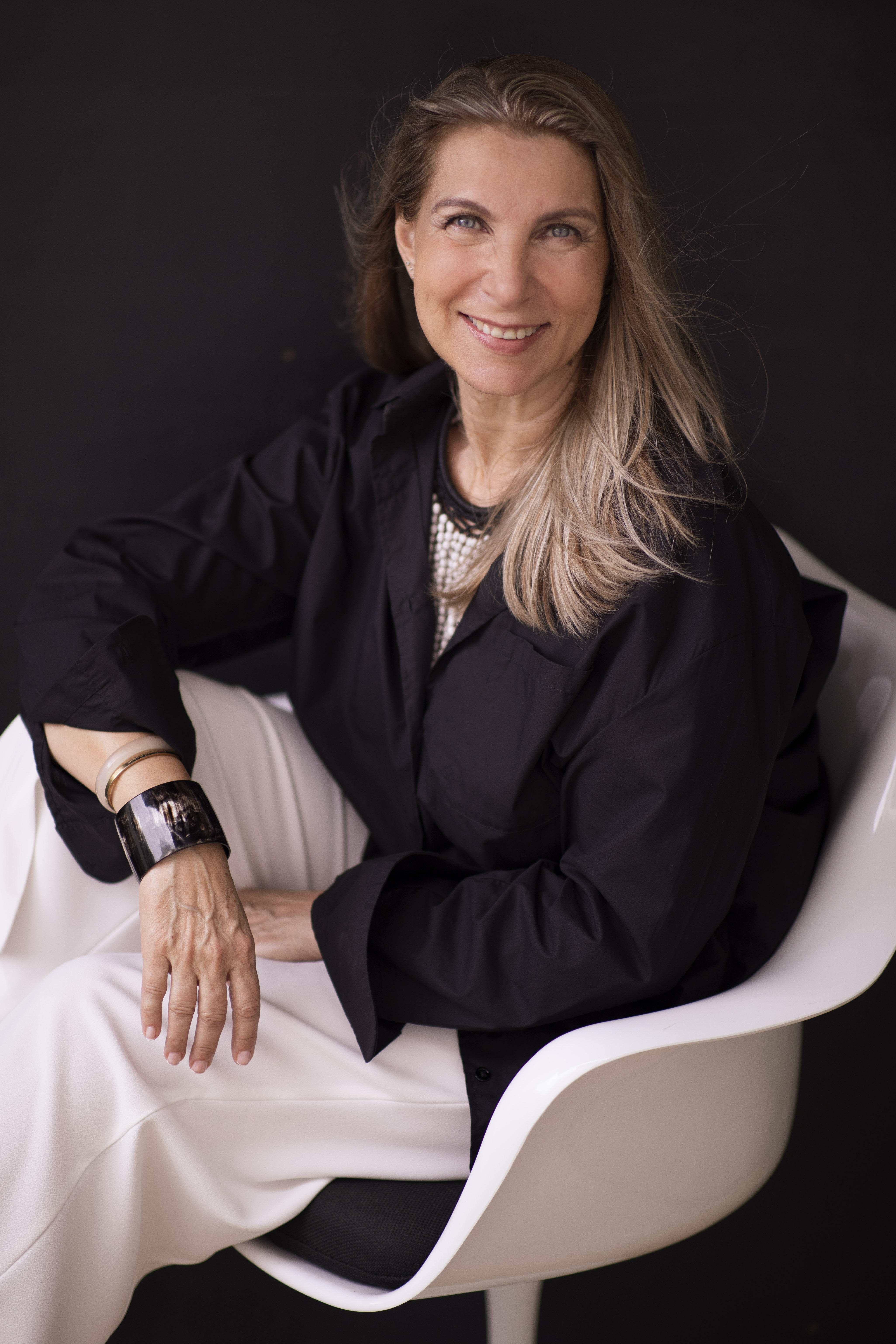 Isabelle Miaja smiles from a white armchair against a dark background, wearing a black shirt and white trousers.