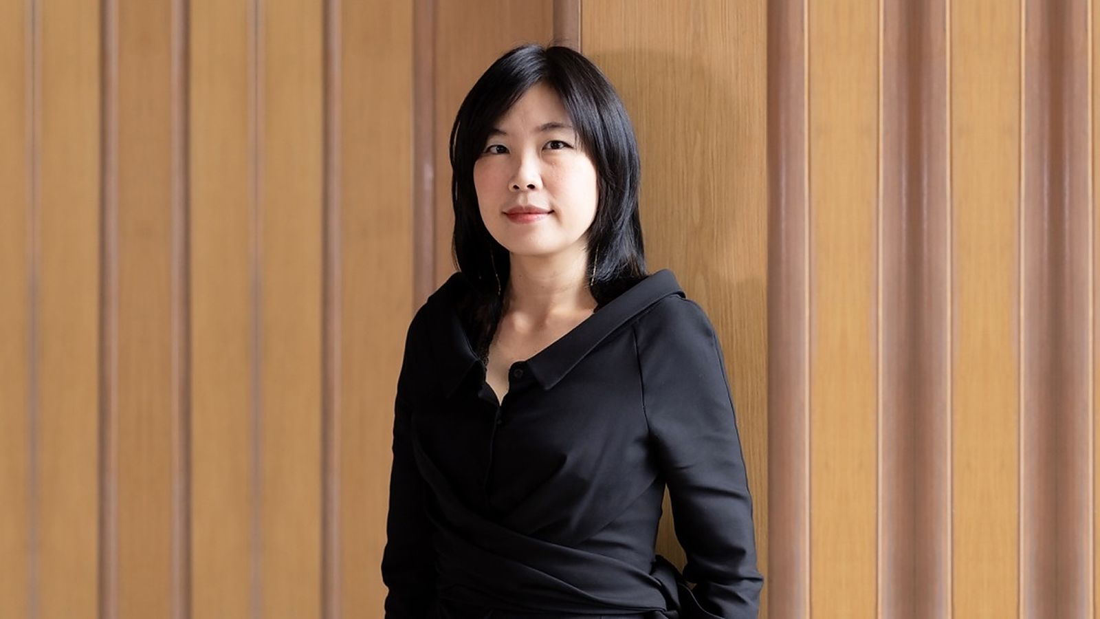 Portrait of Kulthida Songkittipakdee, an architect with shoulder-length black hair wearing a black wrap top, standing against a timber-panelled wall.