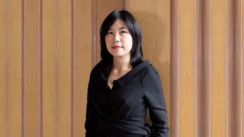 Portrait of Kulthida Songkittipakdee, an architect with shoulder-length black hair wearing a black wrap top, standing against a timber-panelled wall.