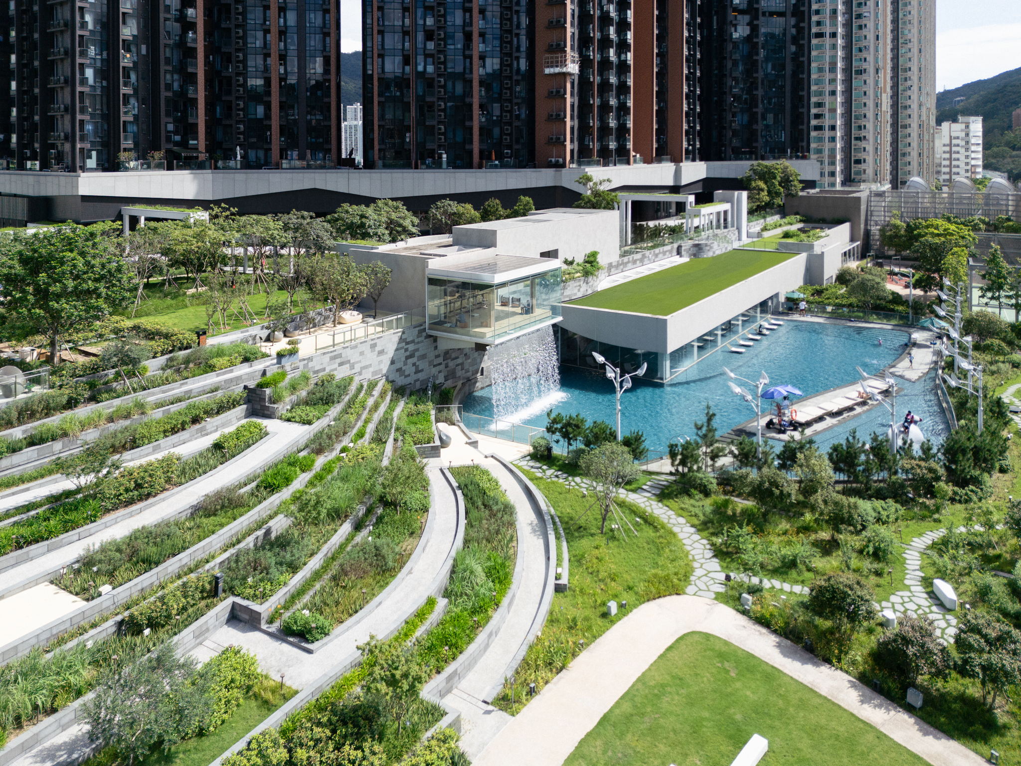 Aerial view of terraced planting beds leading to the Tea House and the Well House, with a long pool and residential towers behind.