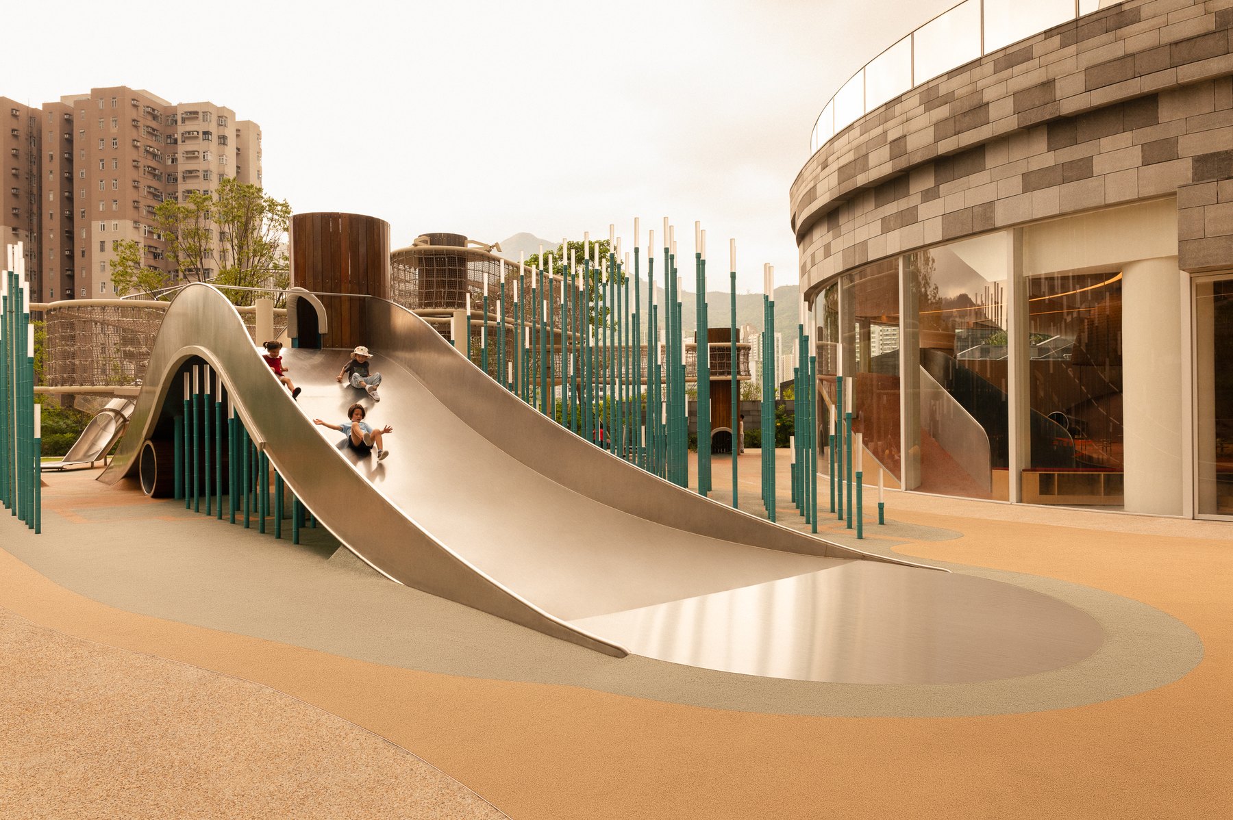A curving stainless steel slide and green vertical posts in the outdoor playground beside the rounded Sky House facade.