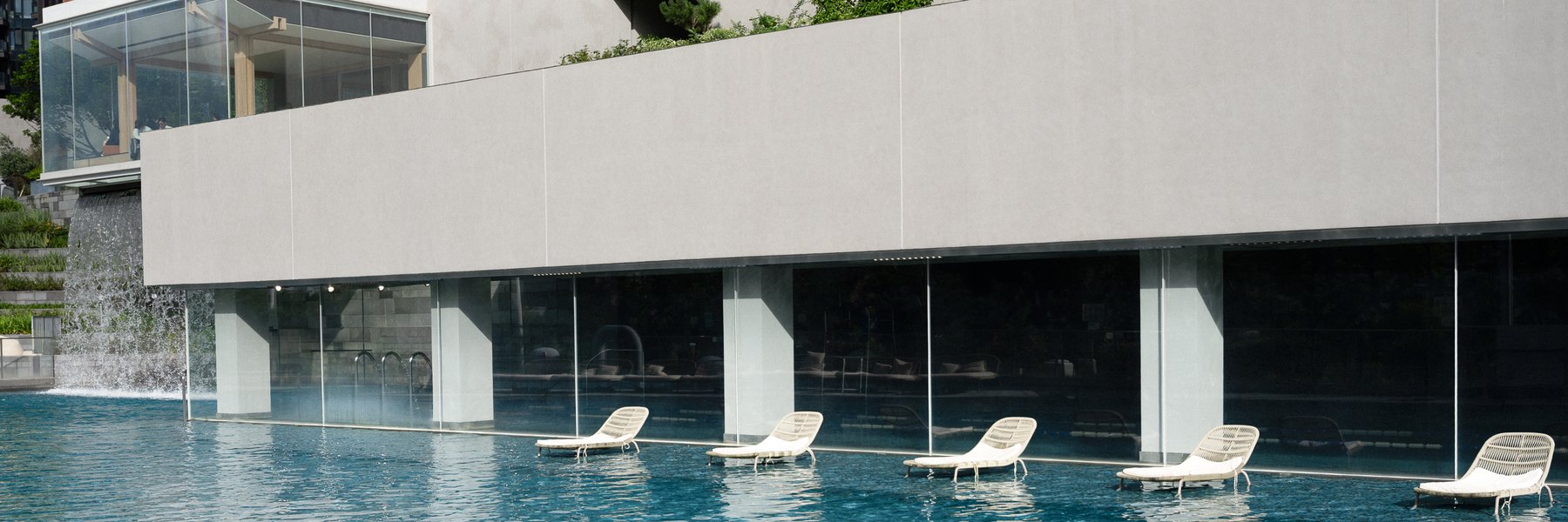 Sun loungers along the edge of the Well House pool, which runs beneath a long grey volume behind full-height glazing.