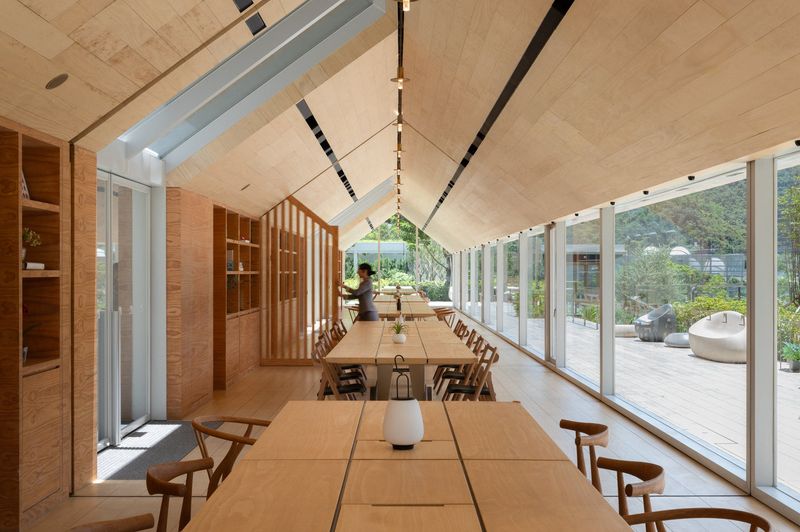 A long gabled dining room lined in pale timber, with rows of wooden tables and chairs beside full-height glazing that opens onto a terrace.